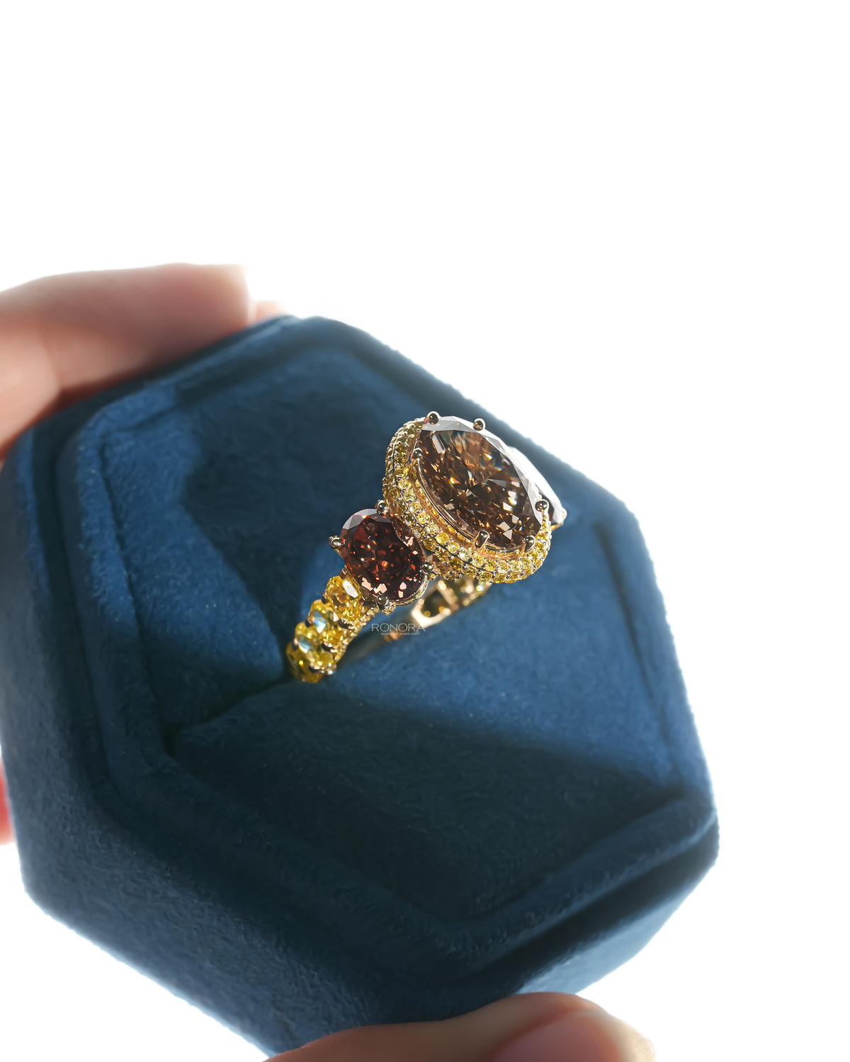 Angled view of a brown oval lab grown diamond three stone ring in a blue velvet box with yellow oval shank stones