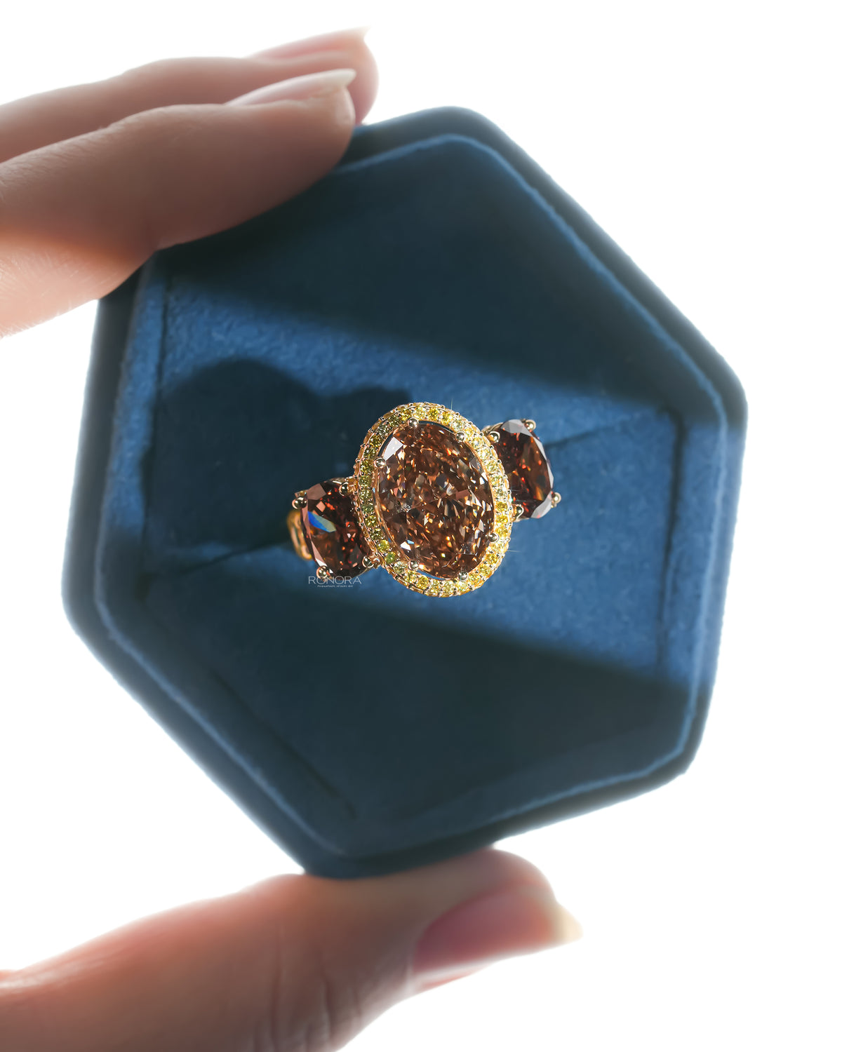Brown oval lab grown diamond three stone ring in a blue velvet hexagon ring box held between two fingers