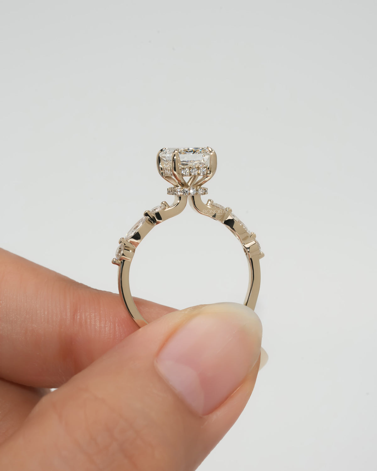 Side view of an emerald cut lab grown diamond ring held up in fingers, showing the slim yellow gold band and side diamonds