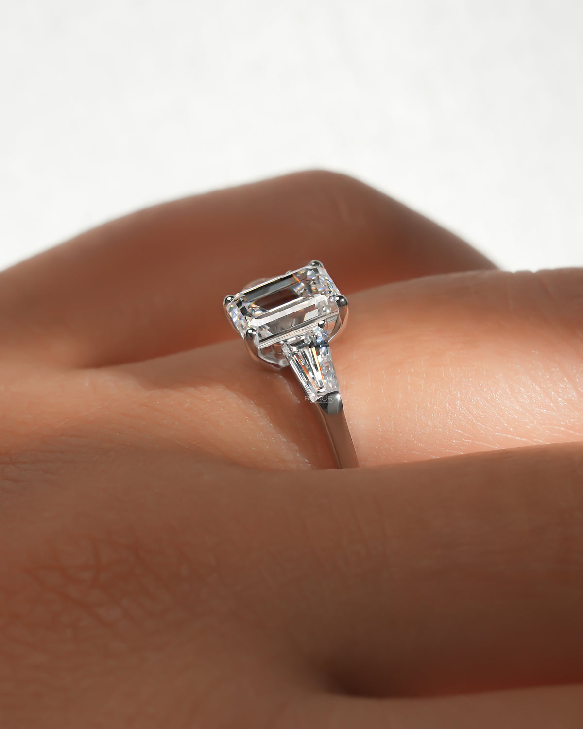 Side view of an emerald cut lab grown diamond three stone ring on a finger, showing prongs and a tapered side stone