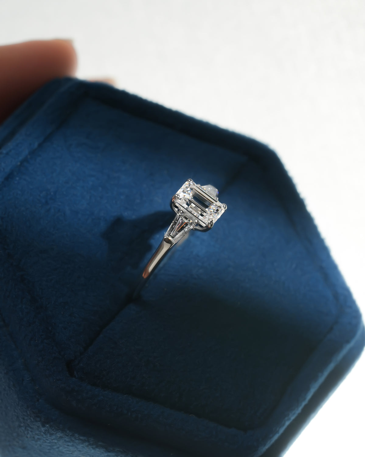 Side angle of an emerald cut lab grown diamond three stone ring in white gold, inside a blue velvet ring box
