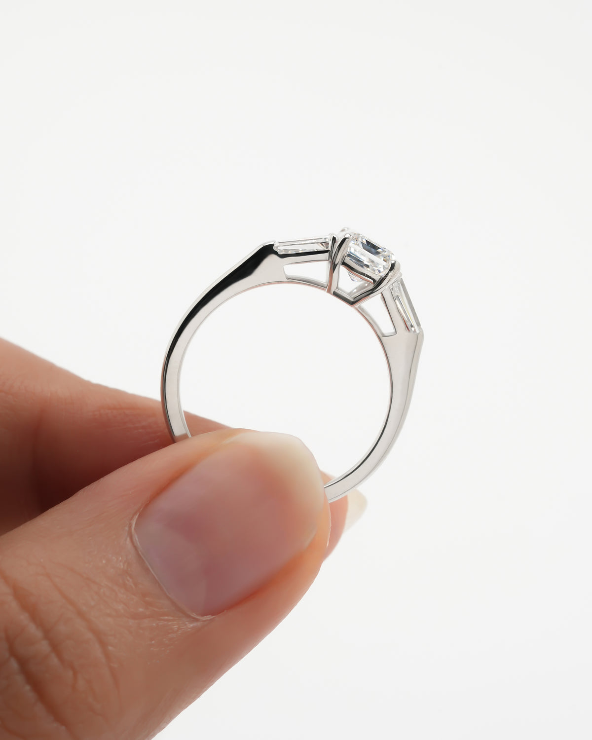 White gold emerald cut lab grown diamond three stone ring held between fingers, side view on a white background