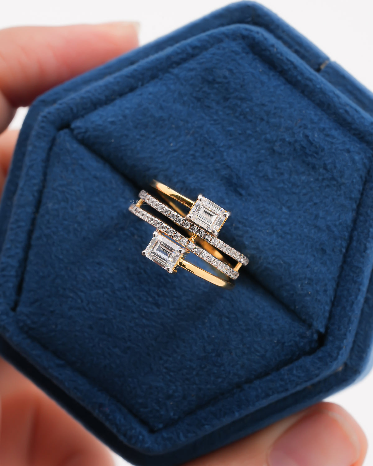 Close-up of an emerald cut toi et moi lab grown diamond ring in yellow gold with pave bands, in a blue velvet box