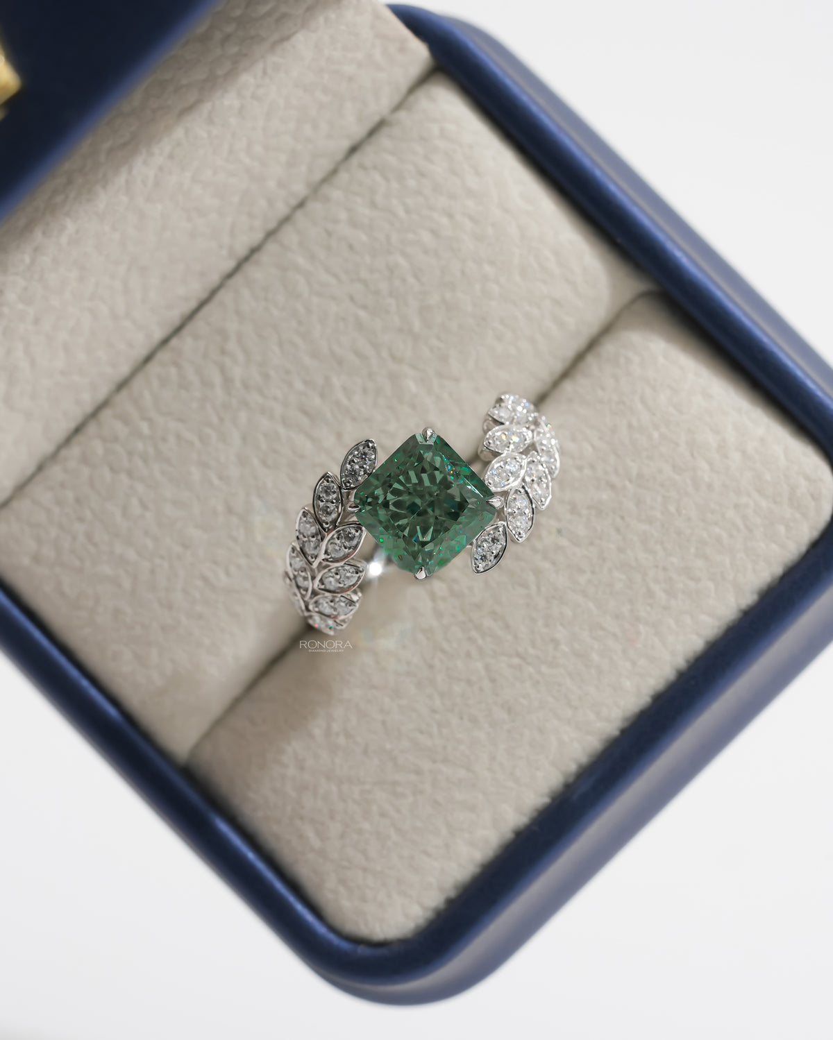 Green cushion lab grown diamond engagement ring with leaf sprig diamond band in a blue ring box, top view