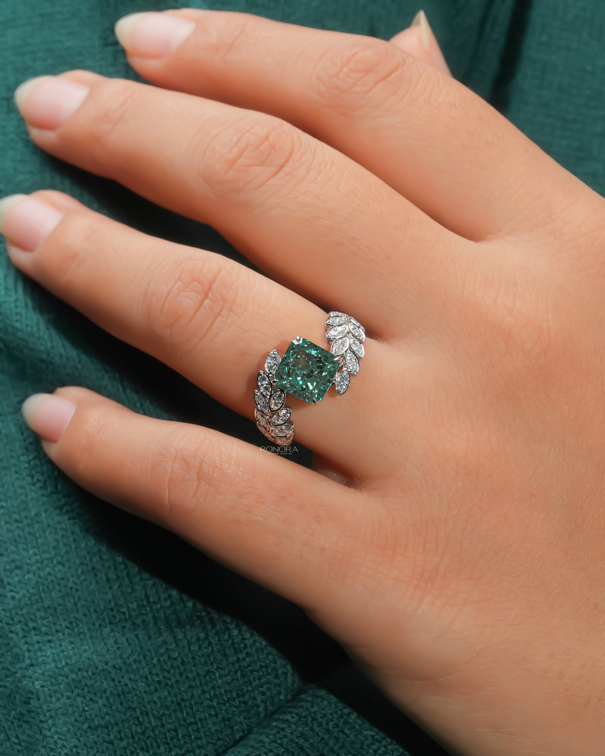 White gold ring with a green cushion lab grown diamond between leaf-shaped shoulders of marquise stones, worn on a hand, green sleeve