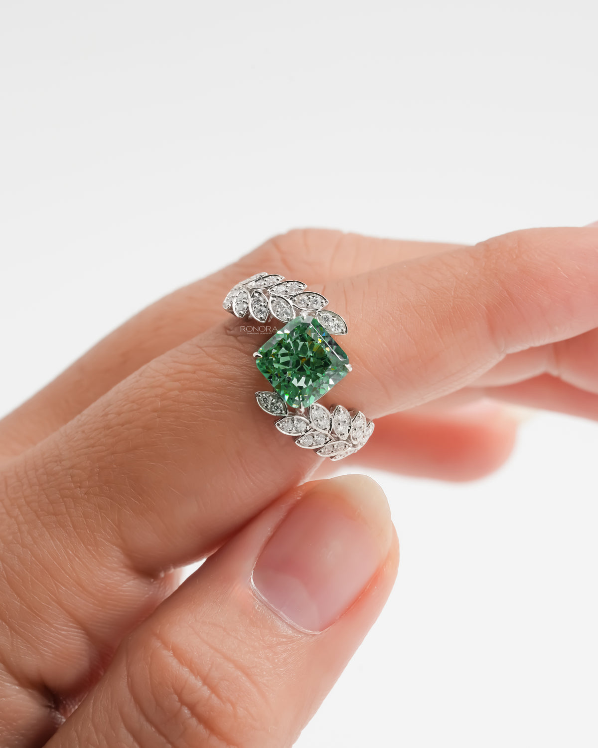Green cushion lab grown diamond engagement ring with leaf sprig diamond bands, held on a finger
