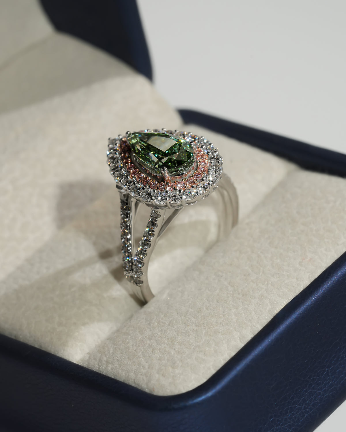 Side angle of a pear green lab grown diamond ring with pink double halo in a cream and blue ring box