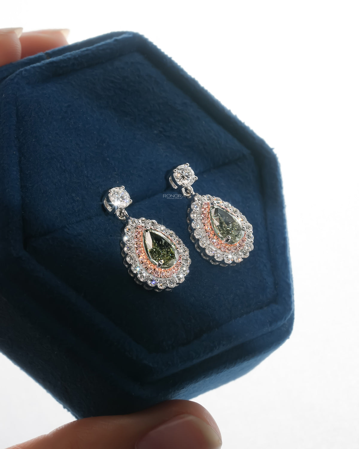 Green pear lab grown diamond drop earrings with pink and white halo resting in a blue velvet jewelry box, angled view