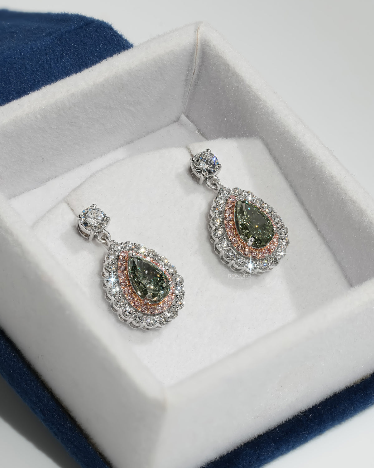 Green pear lab grown diamond drop earrings with pink double halo in a white velvet jewelry box on blue velvet