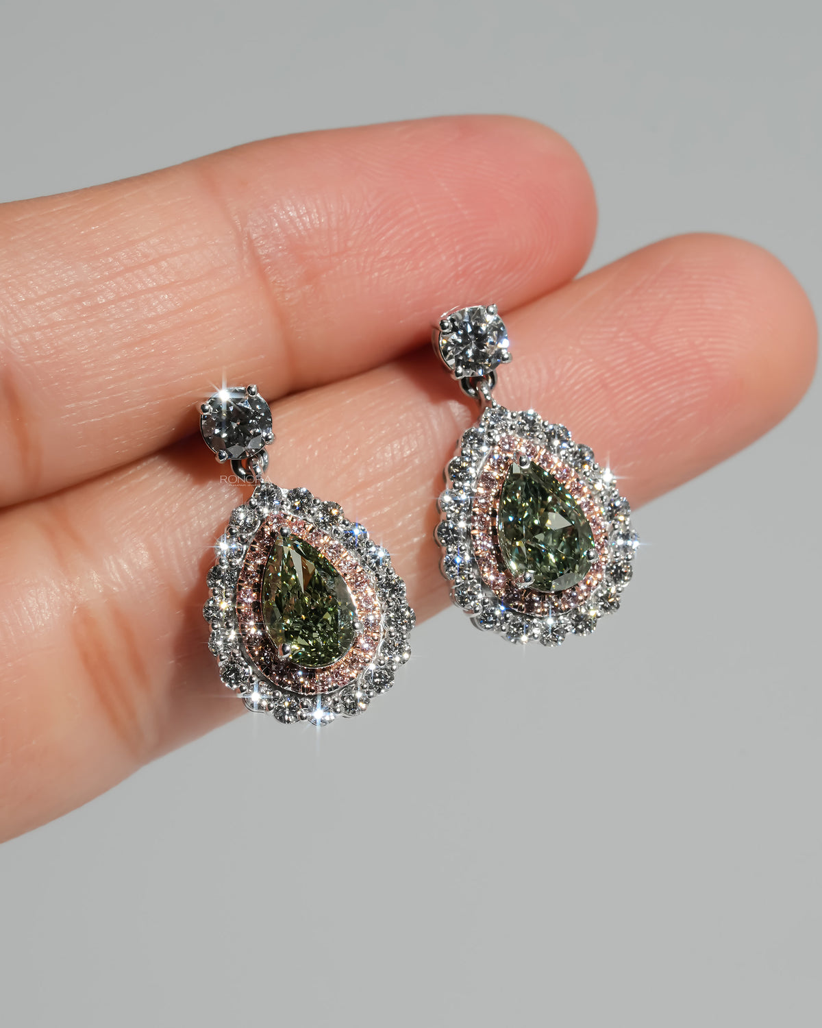 Pair of green pear lab grown diamond drop earrings on fingertips, sparkling pink and white diamond halos in white gold