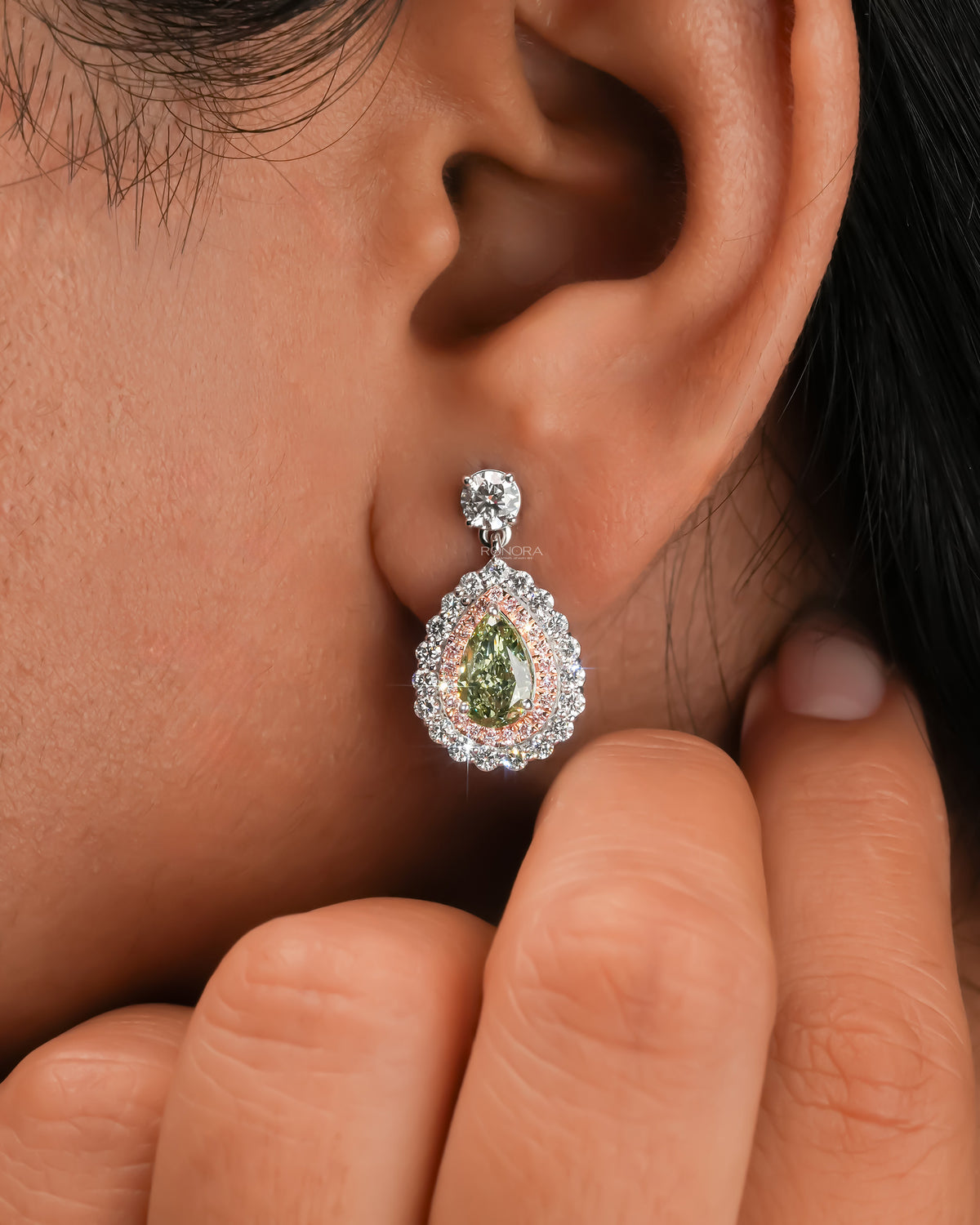 Close-up of an ear wearing a green pear lab grown diamond drop earring with a round diamond top and pink halo