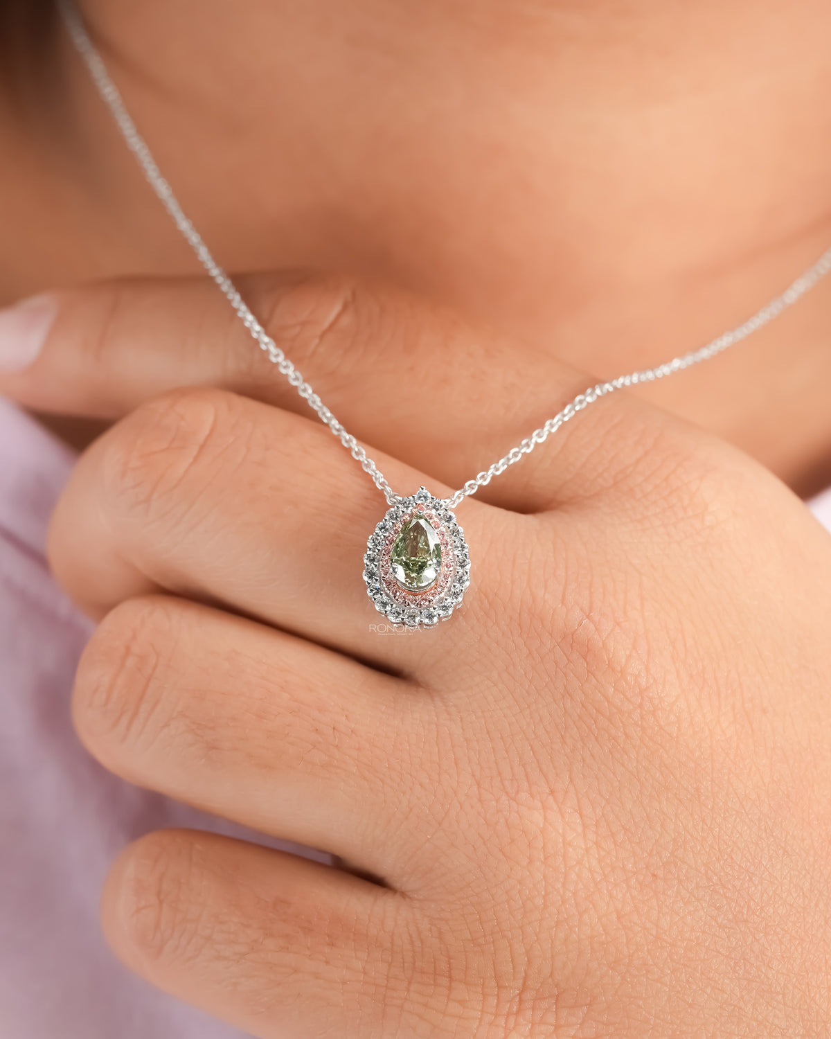 Worn green pear lab grown diamond pendant with a pink double halo, a hand resting near the white gold chain