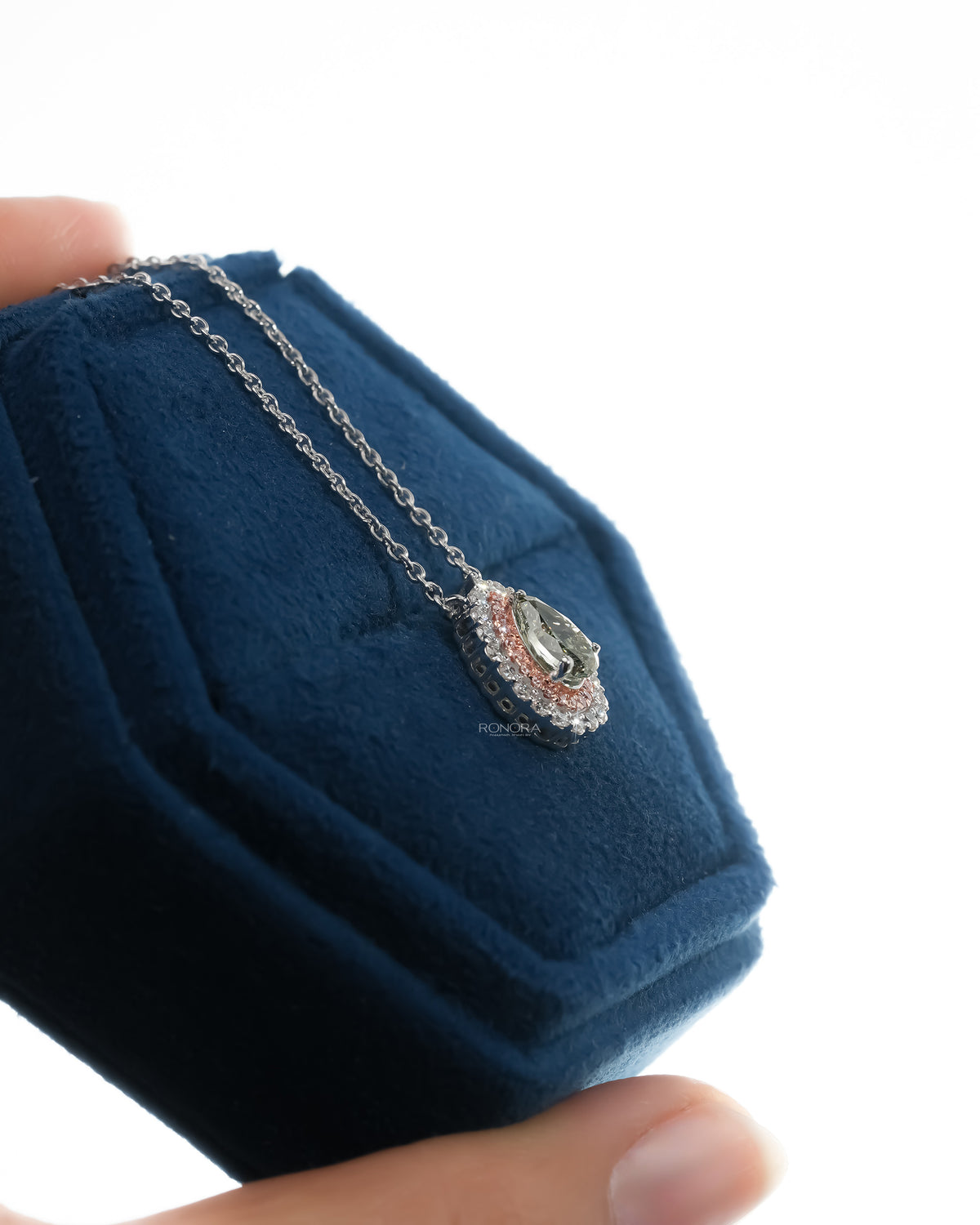Side angle of a green pear lab grown diamond pendant with a pink double halo hanging over a blue velvet box