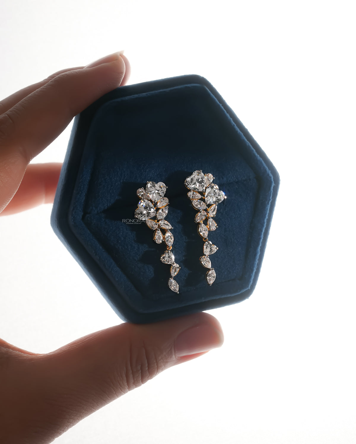 Backlit pair of heart lab grown diamond drop earrings in a blue velvet hexagon box held between fingers
