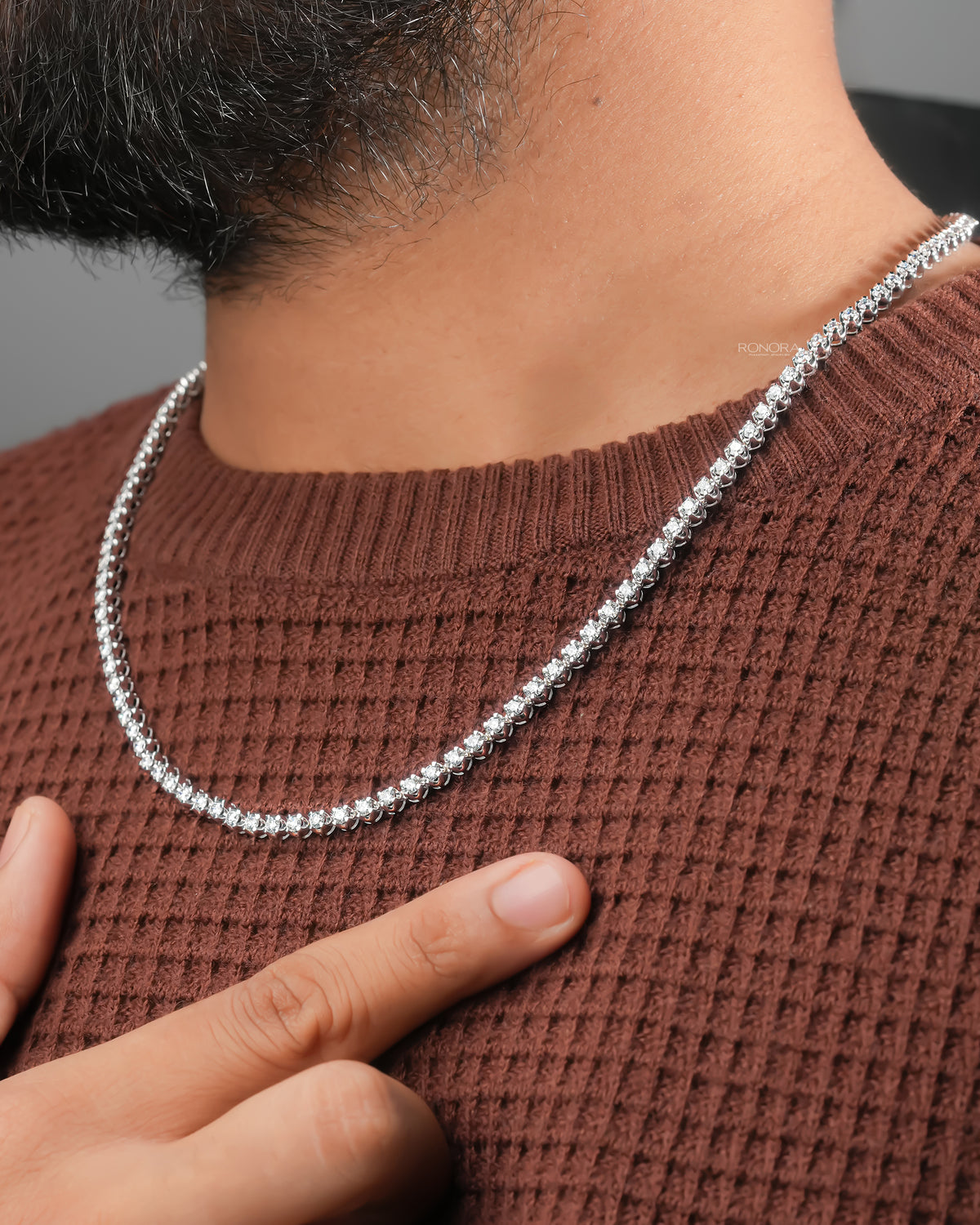 Man's hand pointing to a lab grown diamond tennis necklace worn over a brown knit sweater