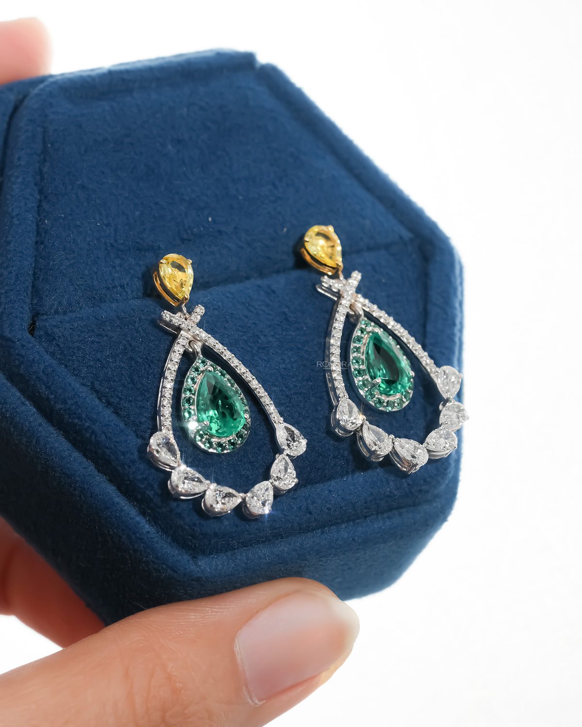 Lab grown emerald pear drop earrings with green halo and yellow pear tops in a blue velvet jewelry box, angled