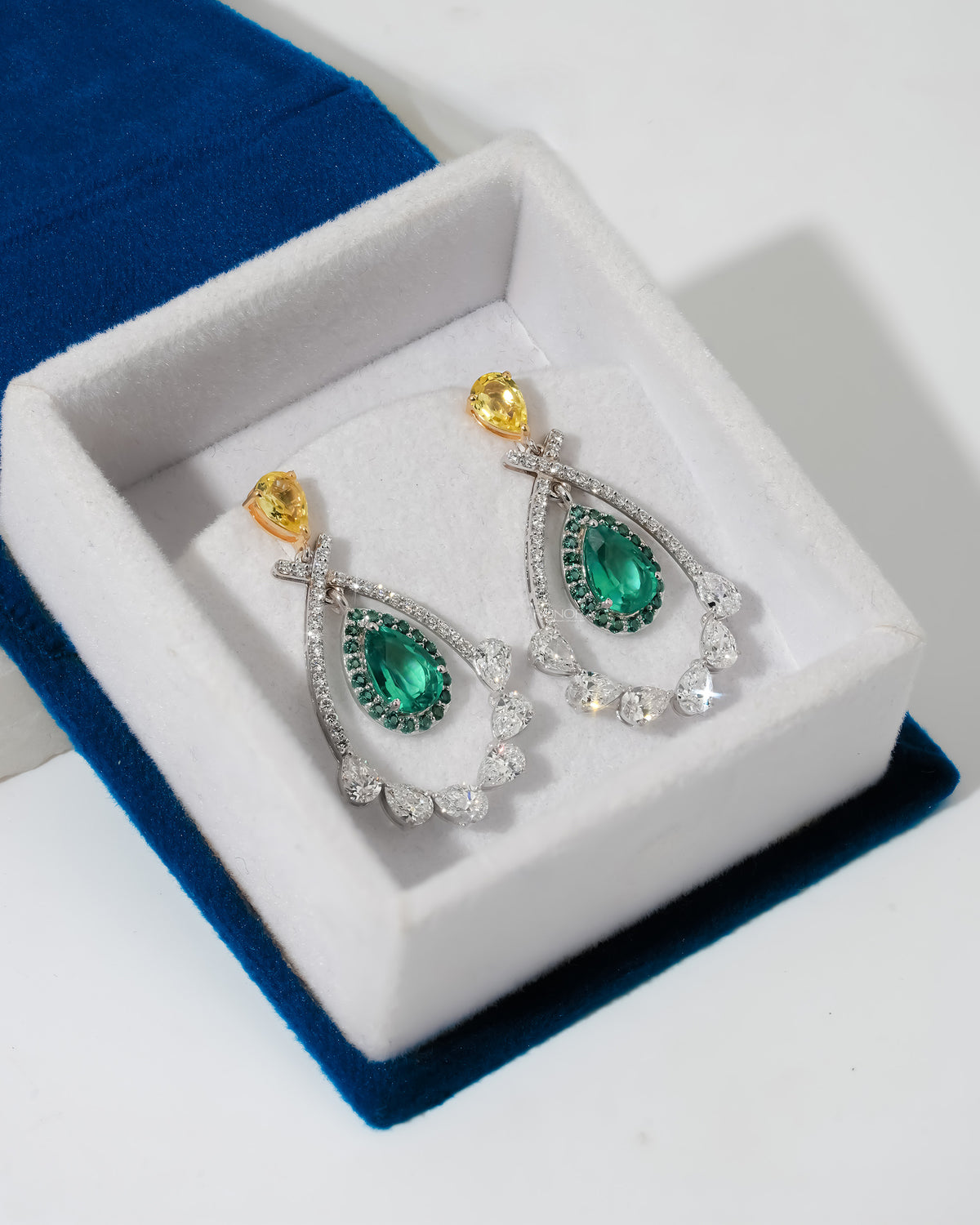 Lab grown emerald pear drop earrings with yellow pear tops in a white velvet jewelry box on blue velvet