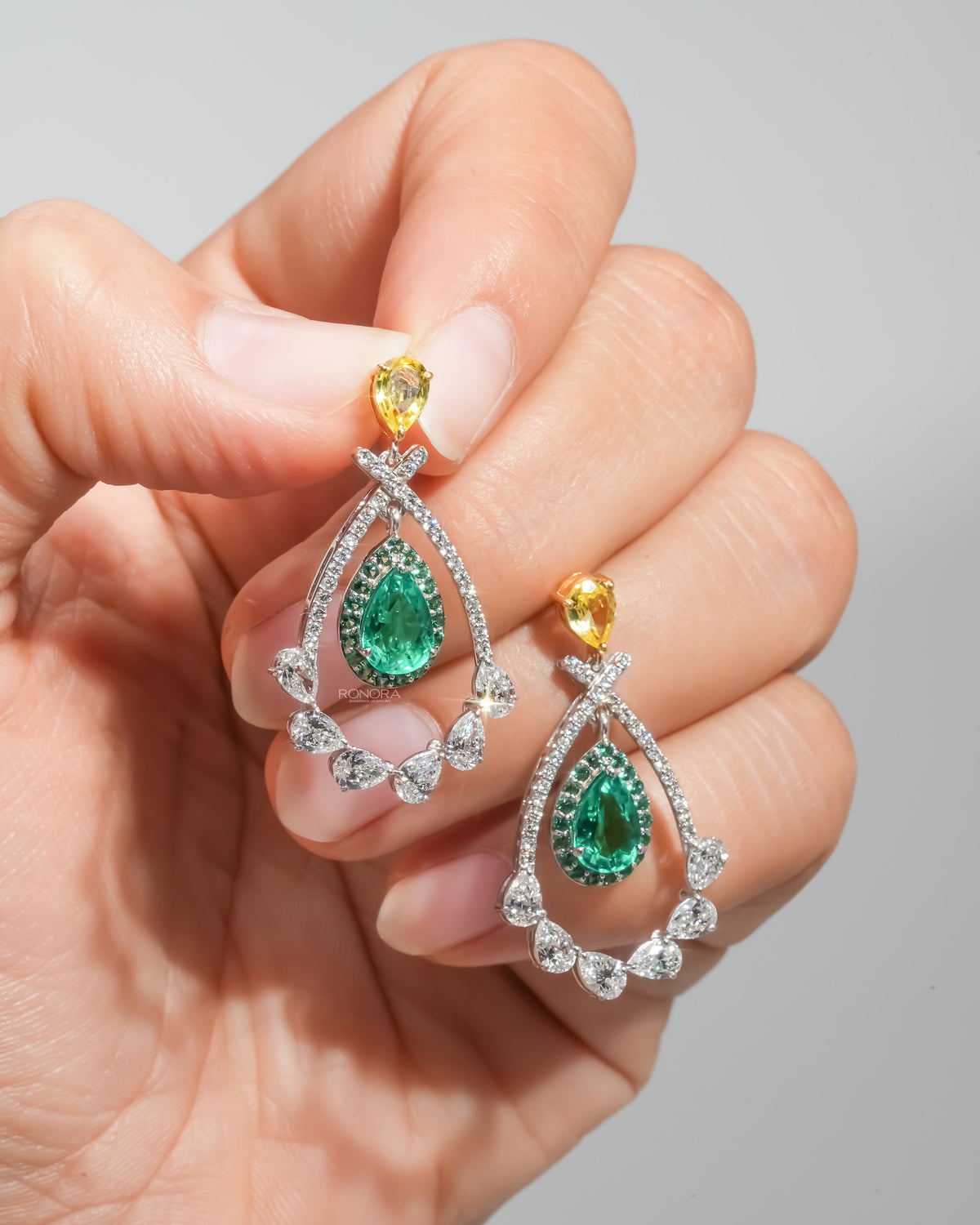 Pair of lab grown emerald pear drop earrings with yellow pear tops held on fingertips, white diamond frame