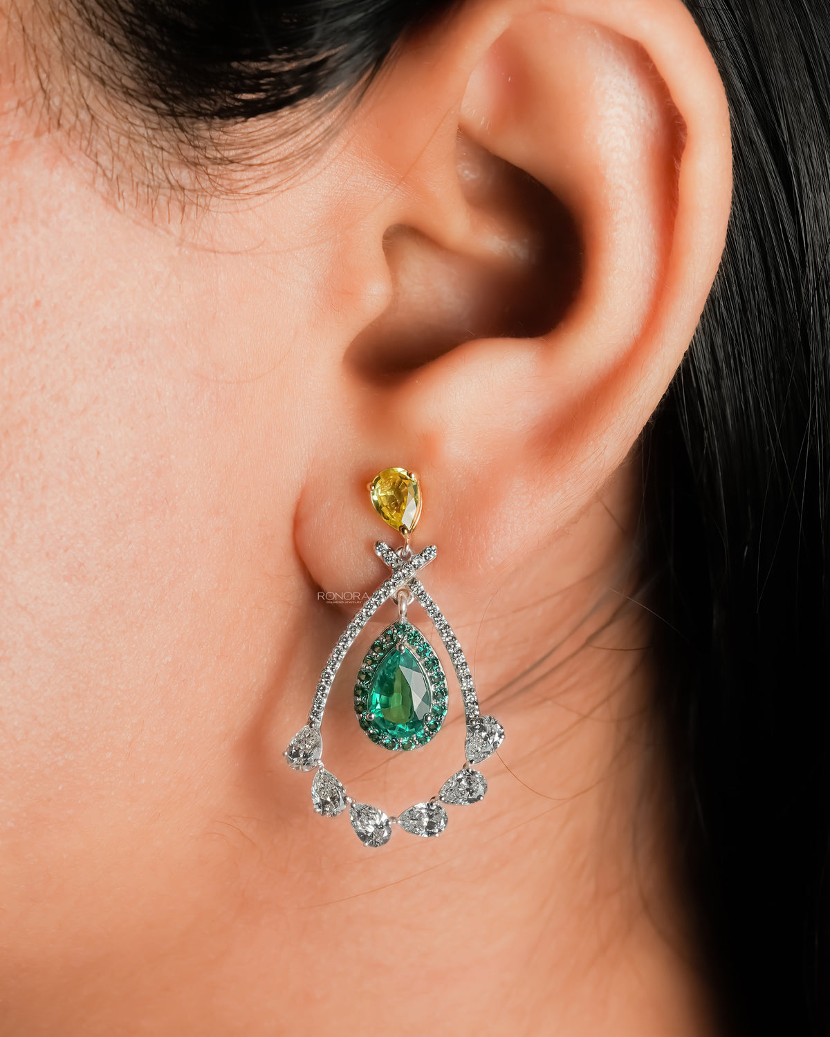 Close-up of an ear wearing a lab grown emerald pear drop earring with green halo and a yellow pear top
