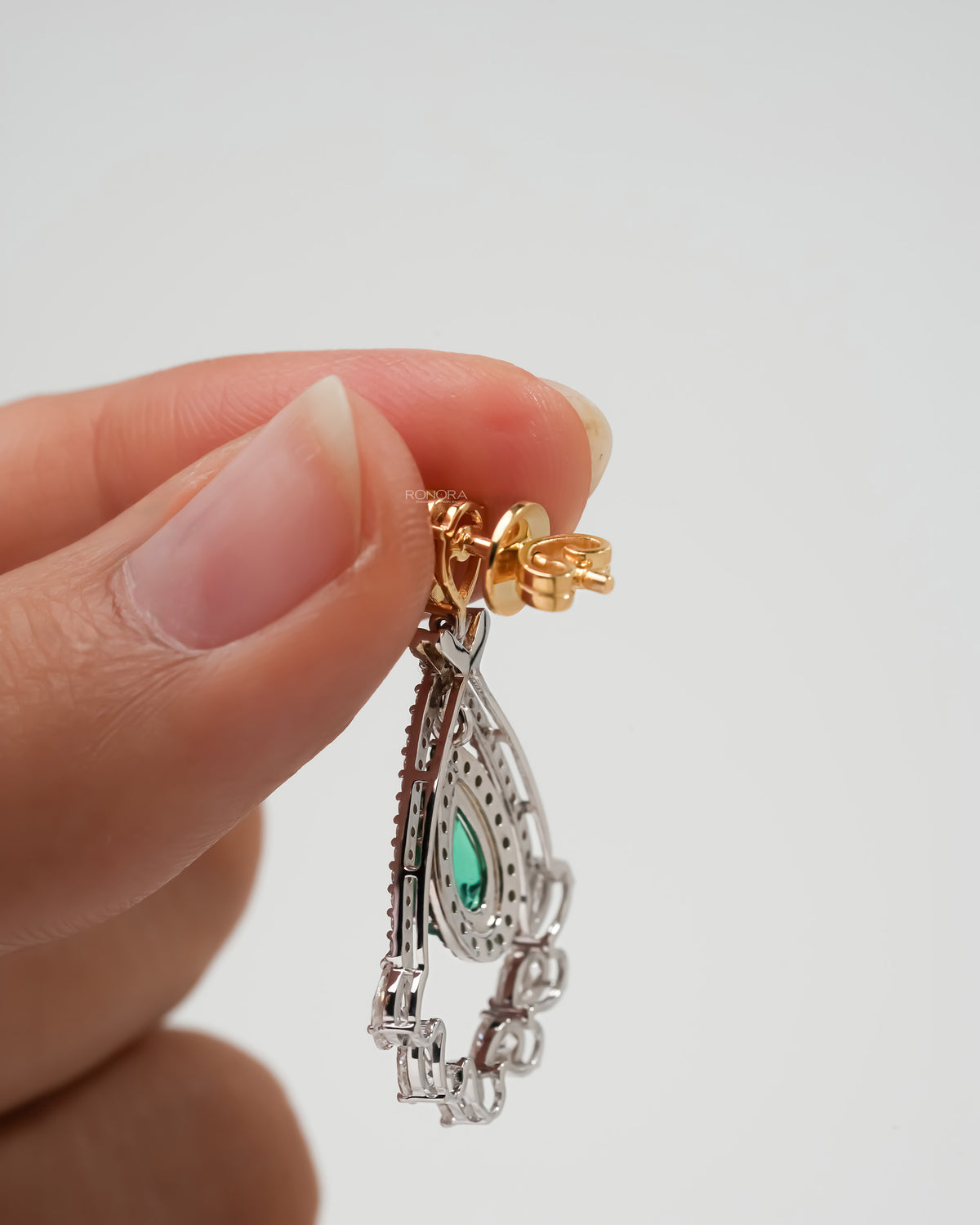 Side view of lab grown emerald pear drop earrings held in fingers, yellow gold post and white diamond frame