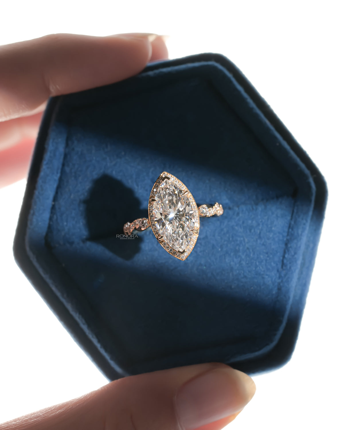 Marquise halo lab grown diamond ring with a twist band in a blue velvet hexagon ring box held between fingers