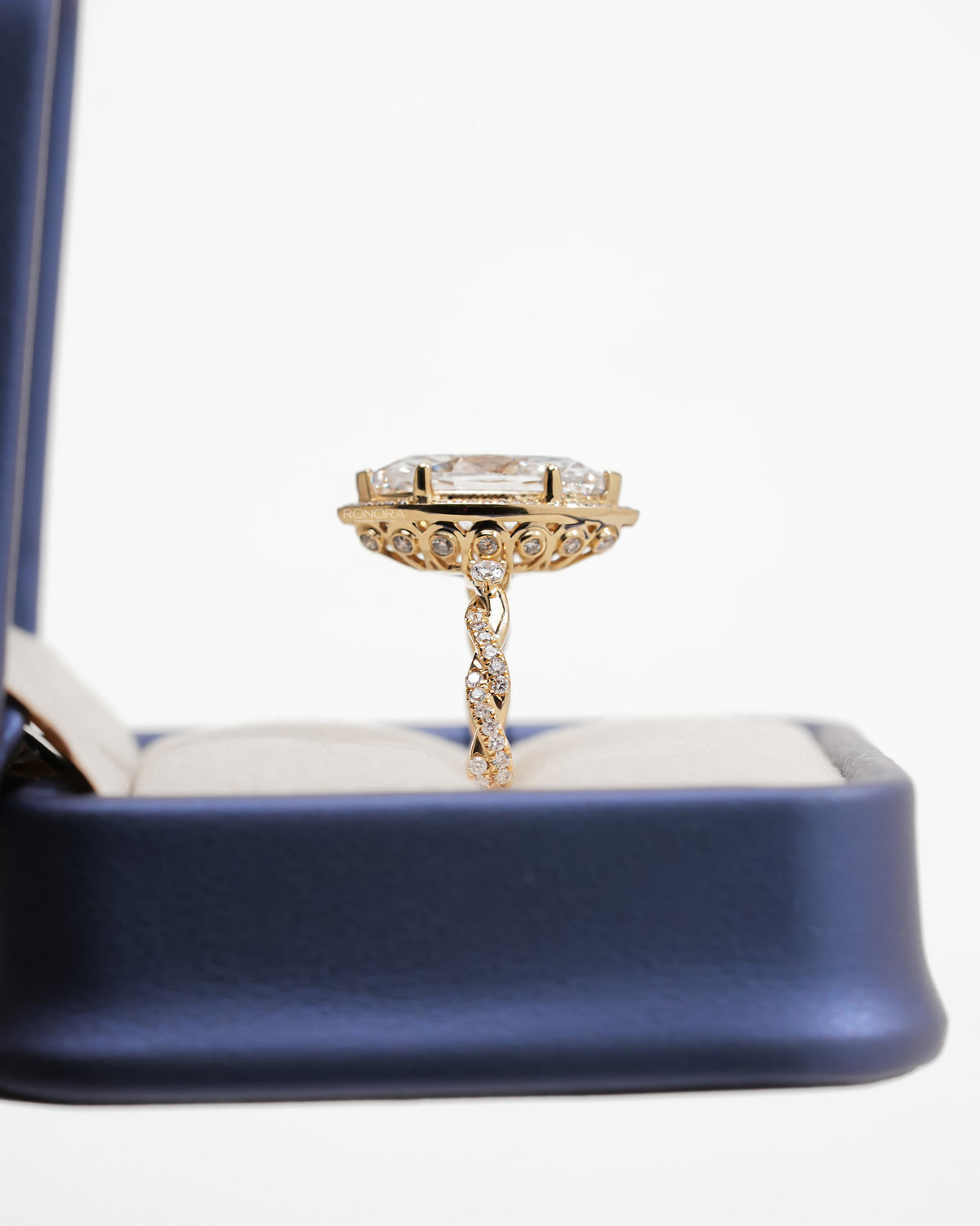 Side view of a marquise halo lab grown diamond ring in yellow gold showing the twisted pave band in a navy box