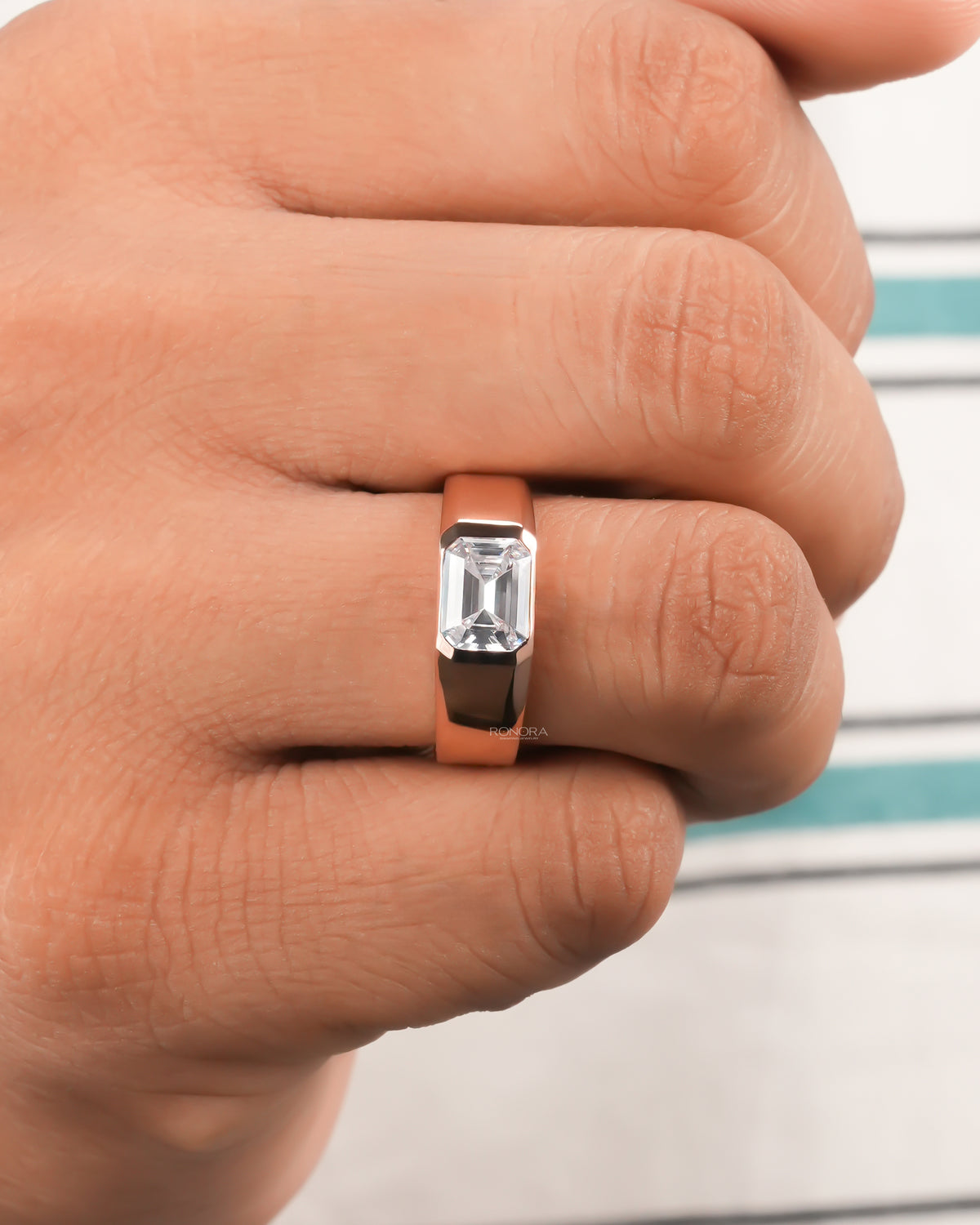 Hand wearing a rose gold men's east west emerald cut lab grown diamond ring with a flush bezel and wide polished band