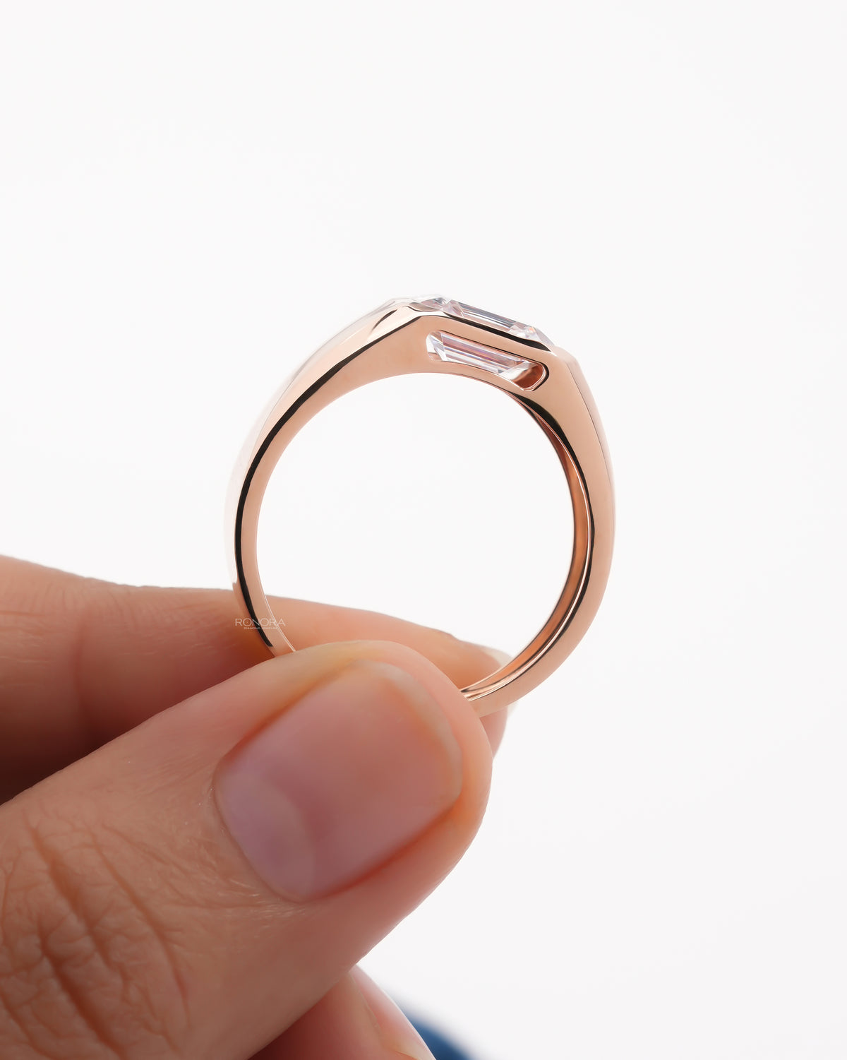 Profile of a rose gold men's emerald cut lab grown diamond ring held between two fingers, flush bezel and wide band
