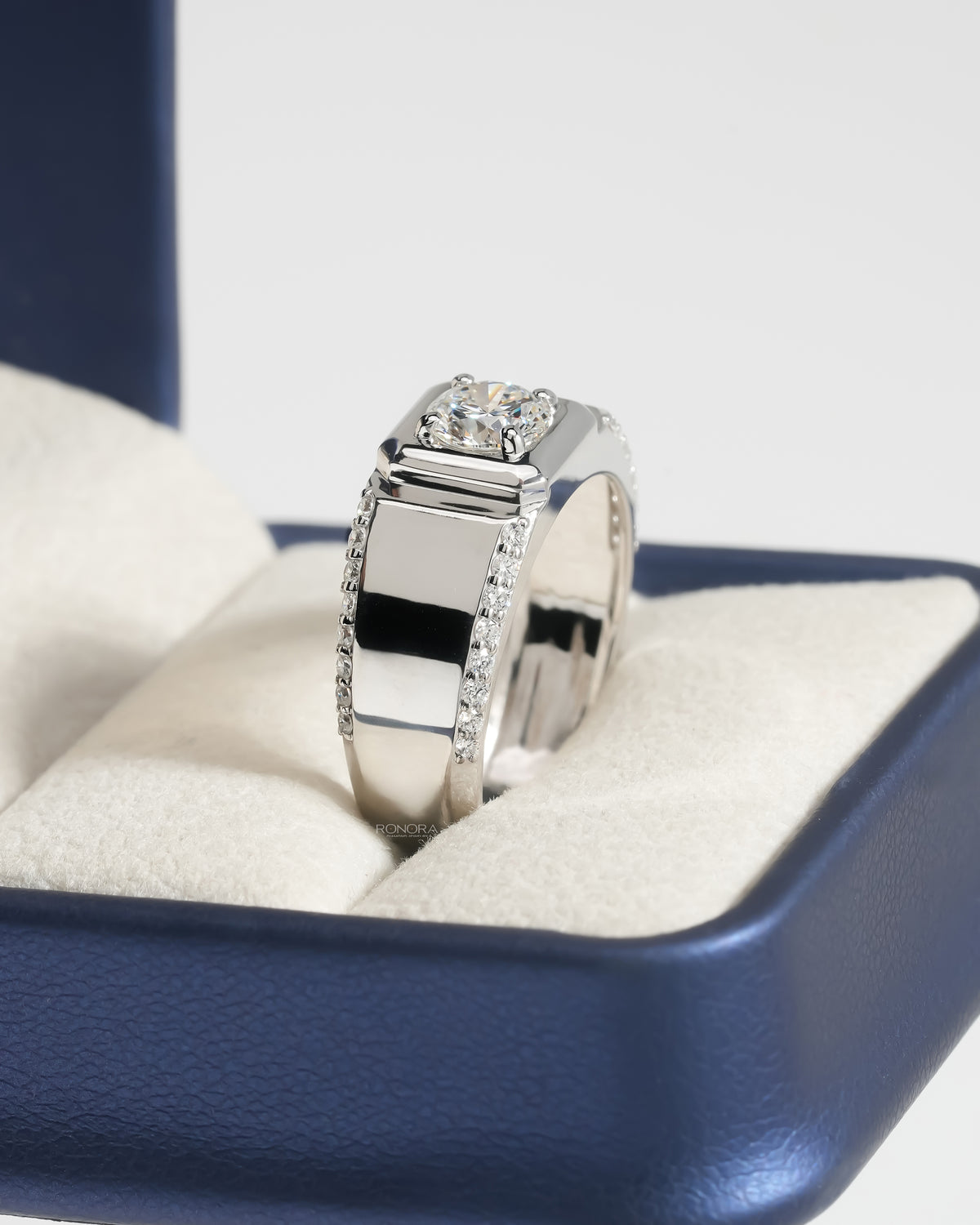 Men's round lab grown diamond ring in white gold with a wide pave band in a navy ring box, angled view