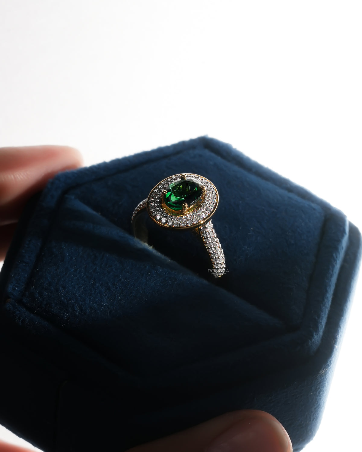 Angled view of an oval green double halo ring with pave band in a blue velvet ring box held in the hand