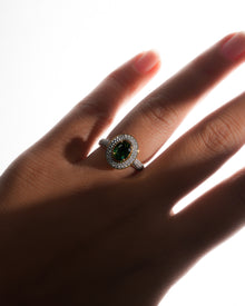 Oval green double halo ring with a pave band worn on a backlit hand, green centre stone and gold bezel
