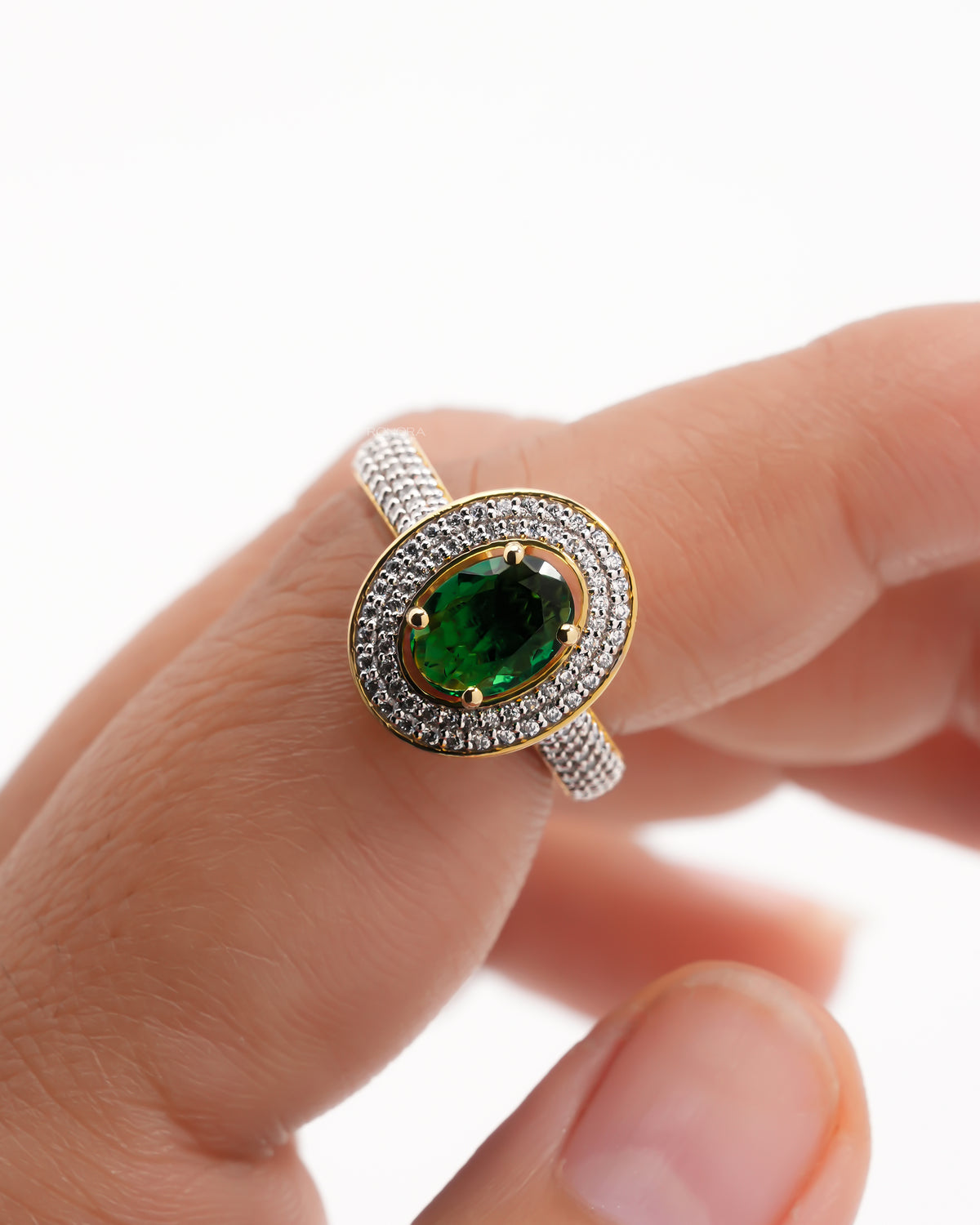 Oval green stone double halo ring in yellow gold with a pave band, held on a finger, close-up front view