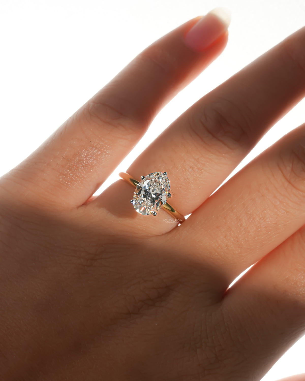 Hand wearing an oval lab grown diamond hidden halo ring with a slim yellow gold band in soft sunlight