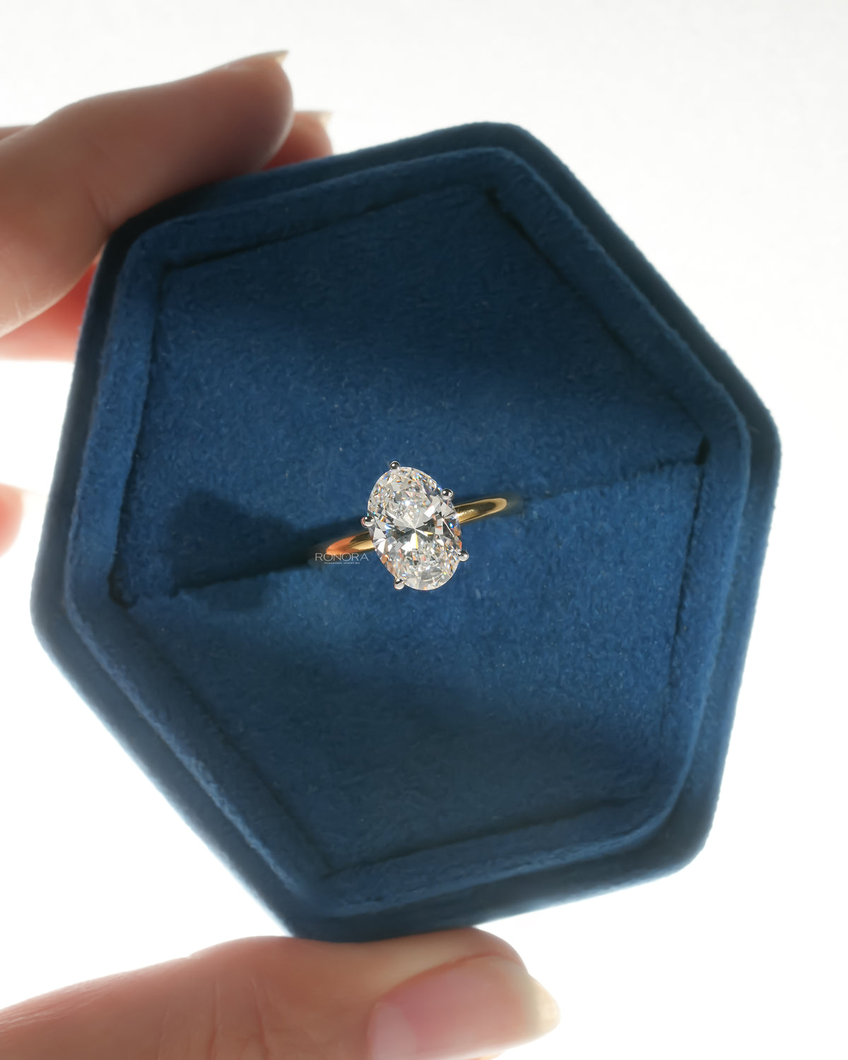 Oval lab grown diamond hidden halo ring in yellow gold in a blue velvet ring box held in fingers, top view