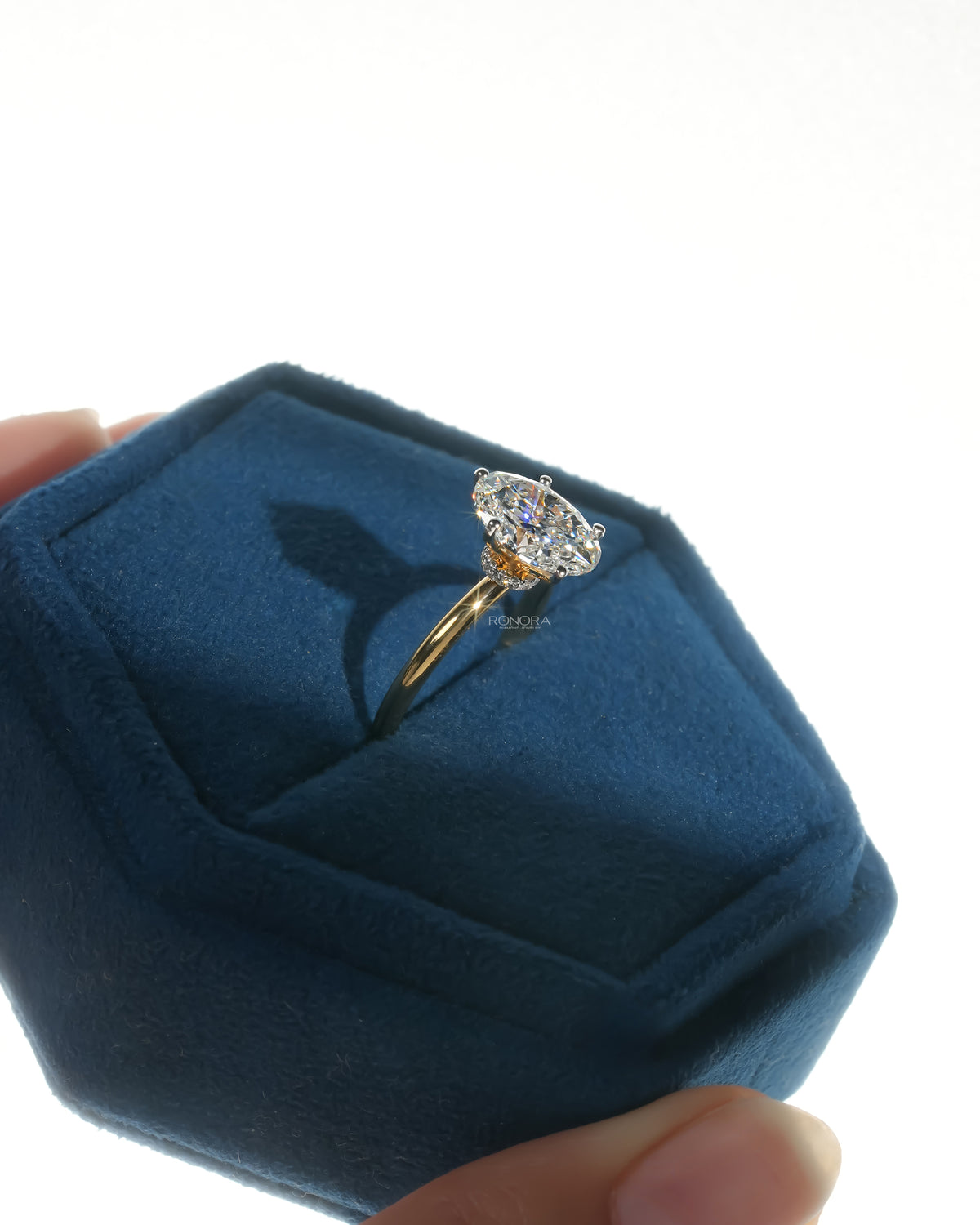 Oval lab grown diamond hidden halo ring in yellow gold inside a blue velvet ring box held in hand