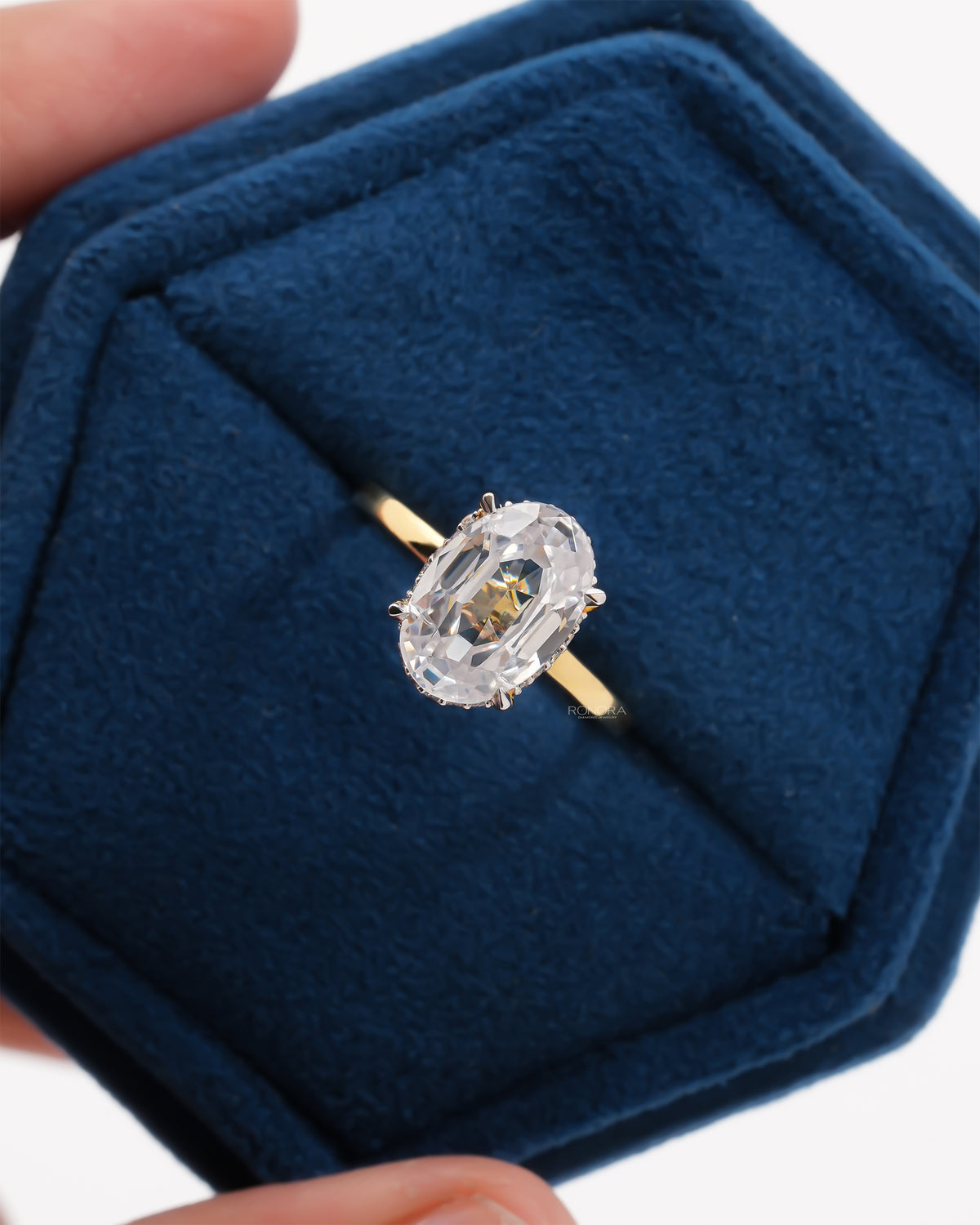 Top view of a 2 carat oval moissanite hidden halo ring with a yellow gold band resting in a blue velvet ring box