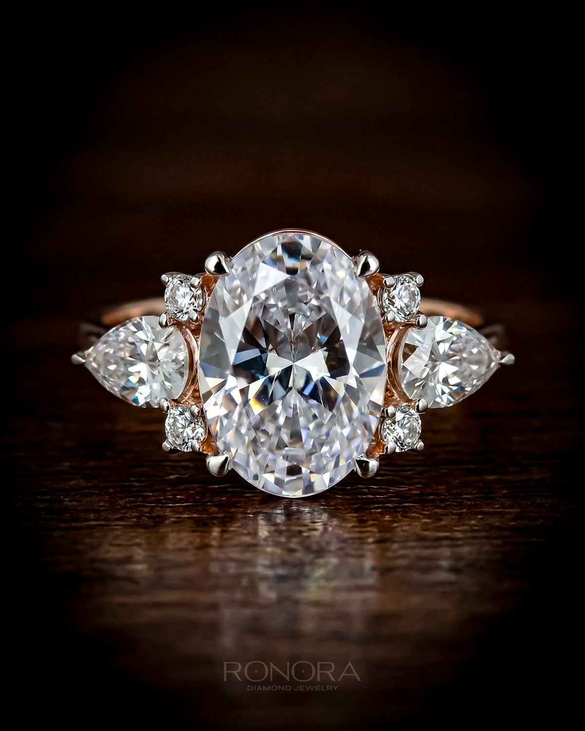 Oval moissanite three stone ring in rose gold with pear side stones and small round accents, close-up on dark wood