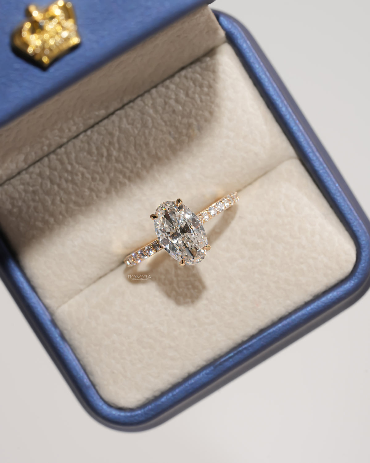 Oval pave lab grown diamond engagement ring in yellow gold in a blue jewelry box with a gold crown on the lid