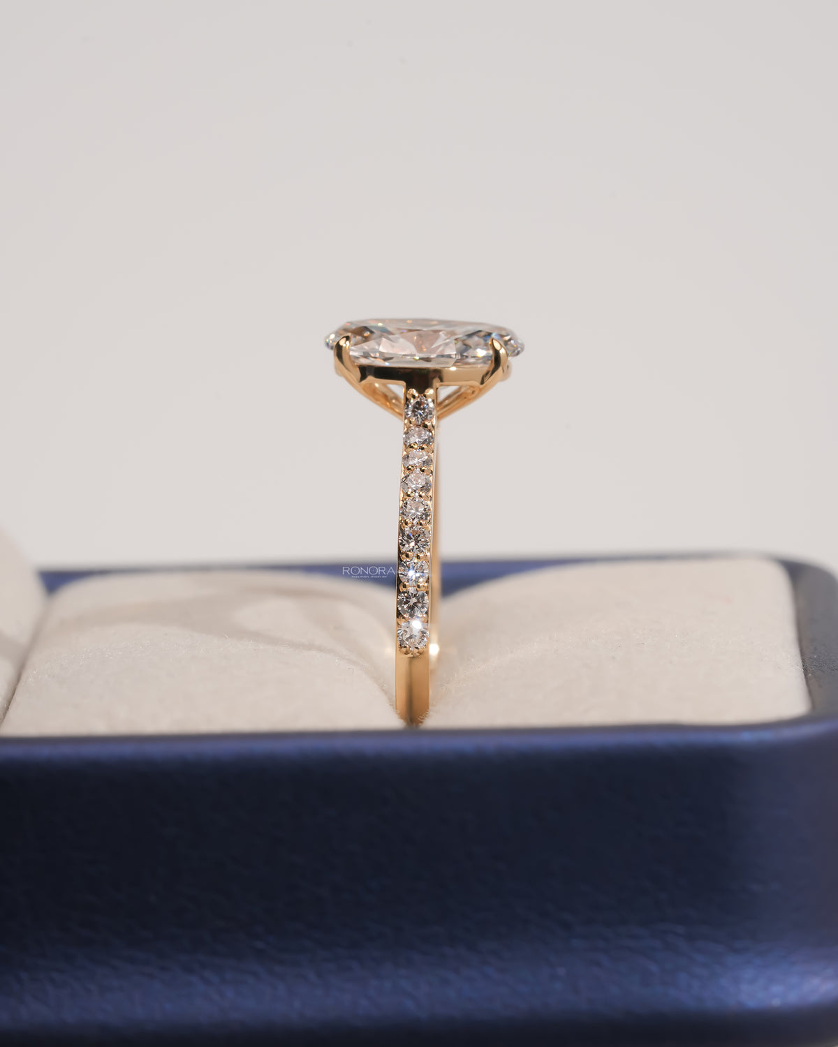 Side view of an oval pave lab grown diamond engagement ring in yellow gold standing upright in a blue ring box
