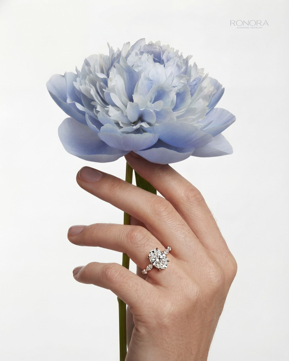 Oval side stone lab grown diamond engagement ring with small side stones, worn on a hand holding a pale blue peony