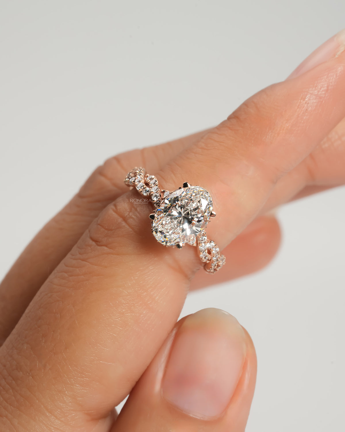 Oval lab grown diamond ring in rose gold with twisted pave band, held between fingertips against a plain grey backdrop