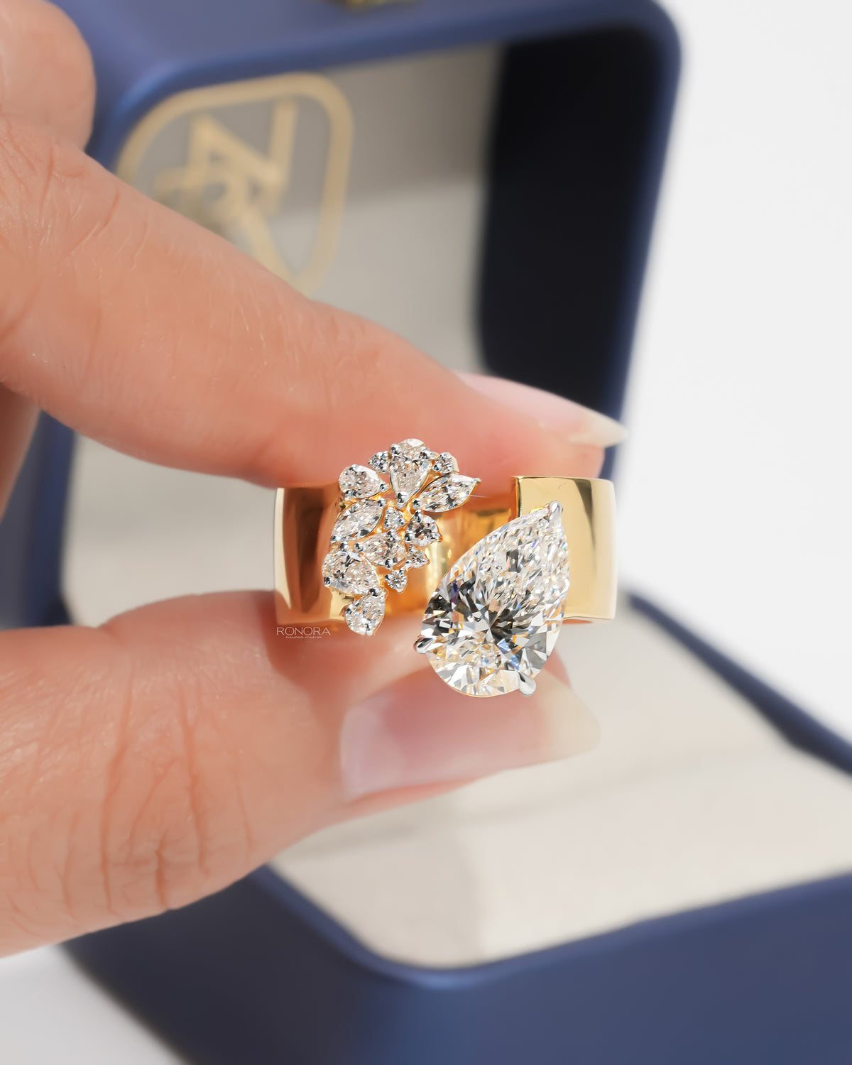 Close-up of a pear toi et moi lab grown diamond ring in yellow gold with a diamond cluster, held in fingers by a ring box