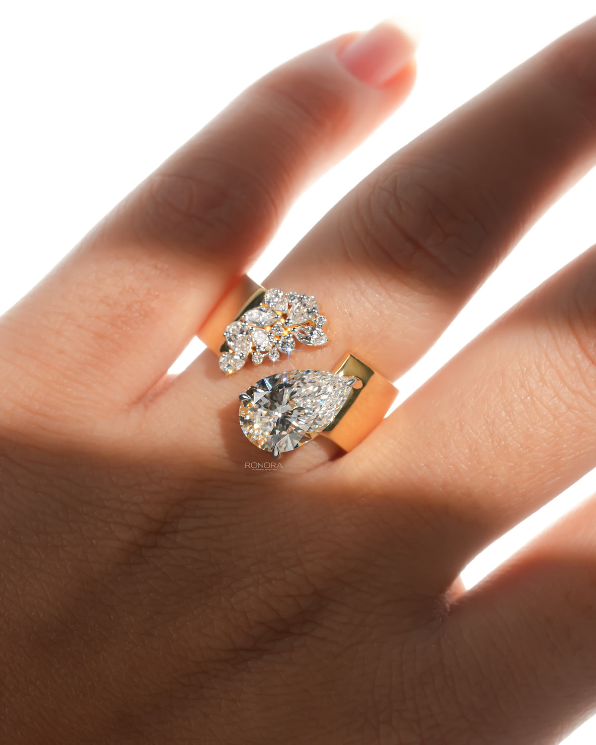 Hand in sunlight wearing a pear toi et moi lab grown diamond ring with an open yellow gold band and cluster