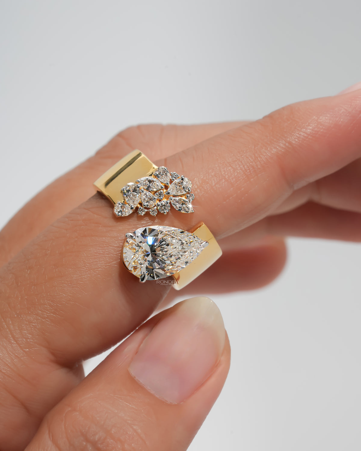 Yellow gold open ring with a pear lab grown diamond at one end and a cluster of marquise and round stones at the other, held in two fingers