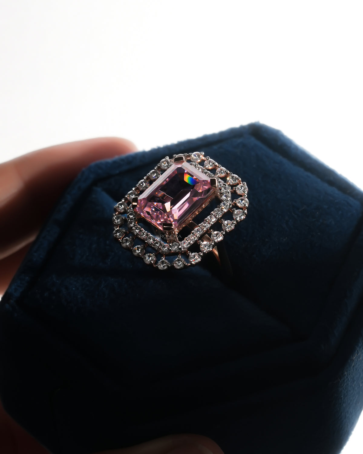 Angled view of a pink emerald cut double halo ring in rose gold sitting in a blue velvet ring box
