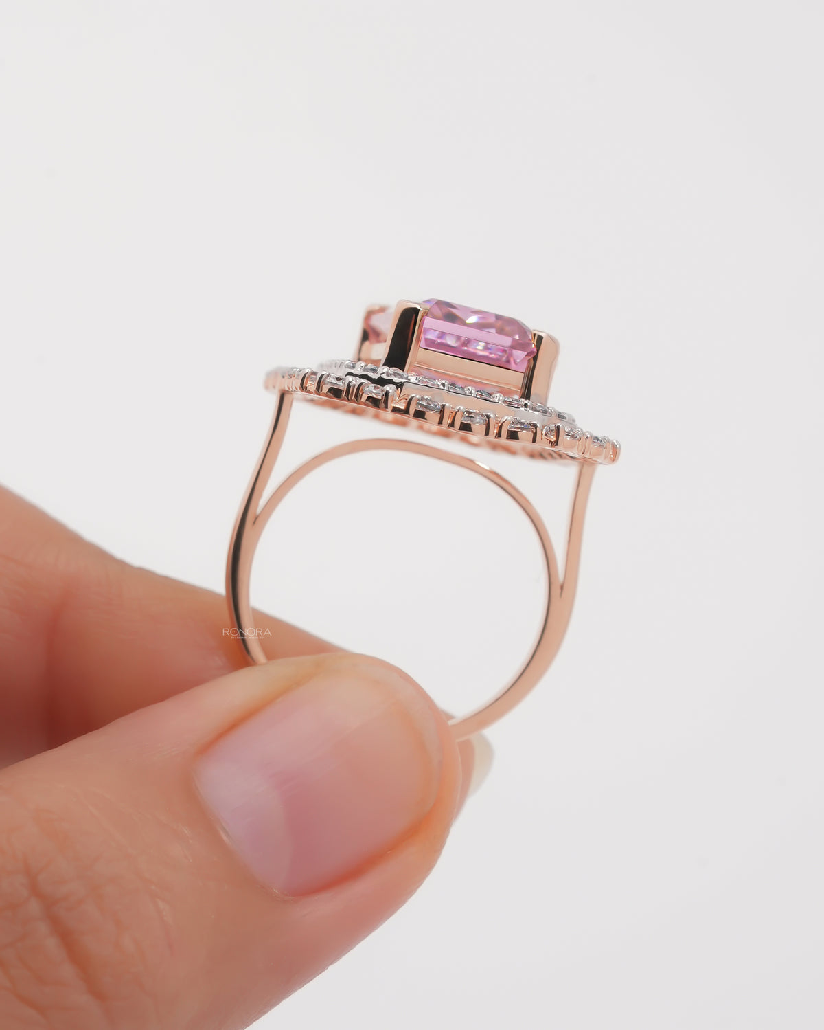 Side profile of a rose gold pink emerald cut double halo ring held upright between two fingers