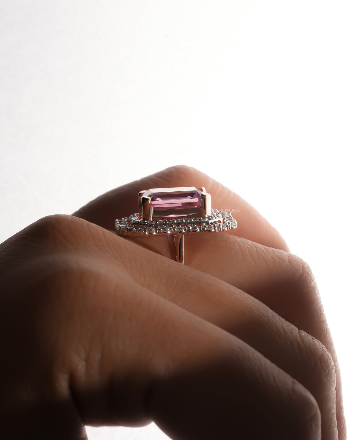 Side view of a pink emerald cut double halo ring in rose gold worn on a finger, backlit
