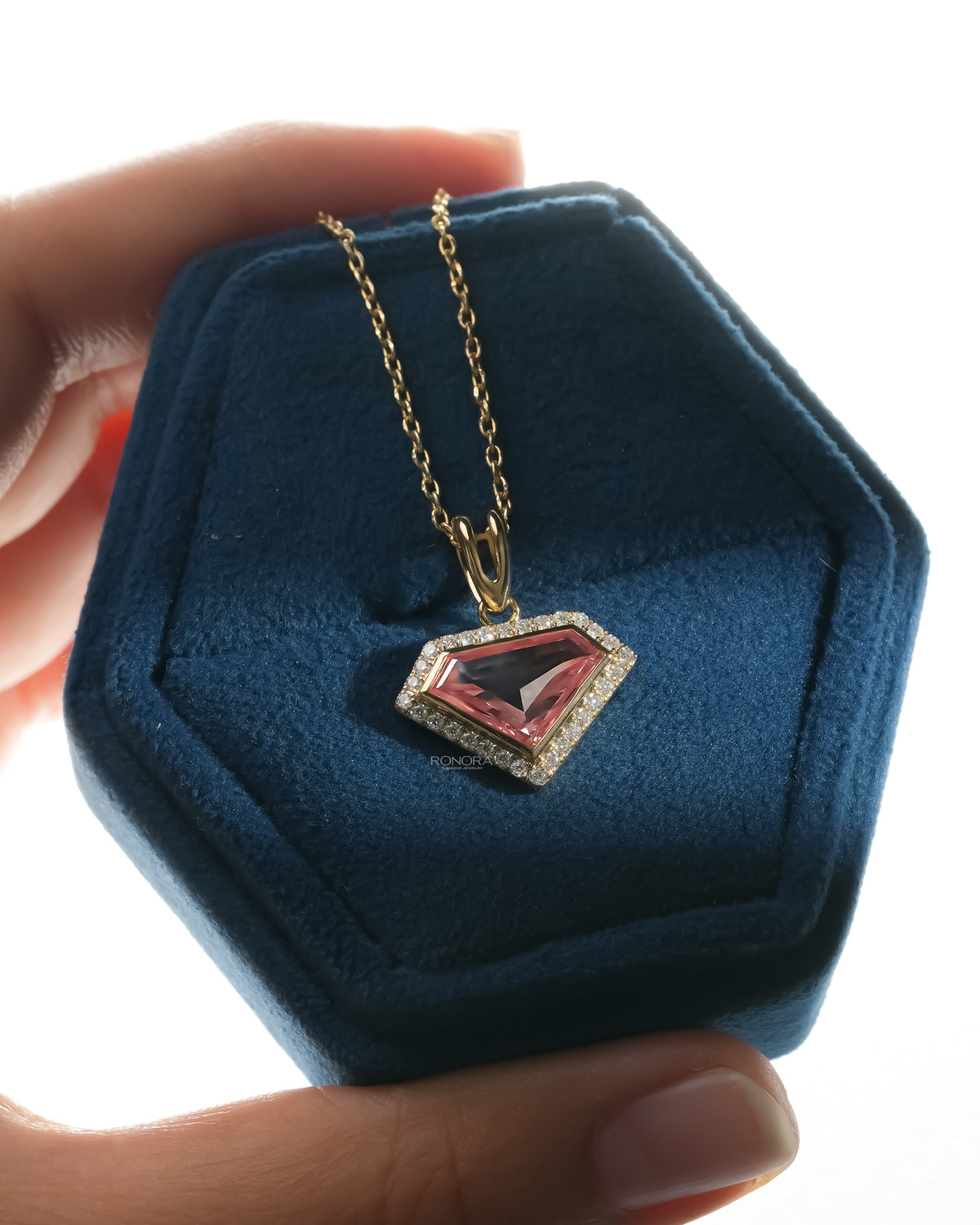 Hand holding a blue velvet hexagon jewelry box with a pink shield cut lab grown diamond necklace in yellow gold