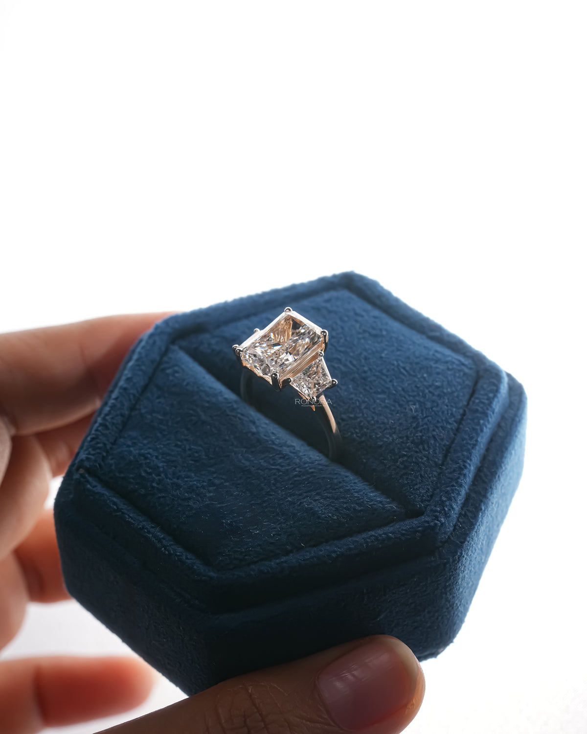 Radiant cut lab grown diamond three stone ring in a blue velvet hexagon jewelry box held in hand