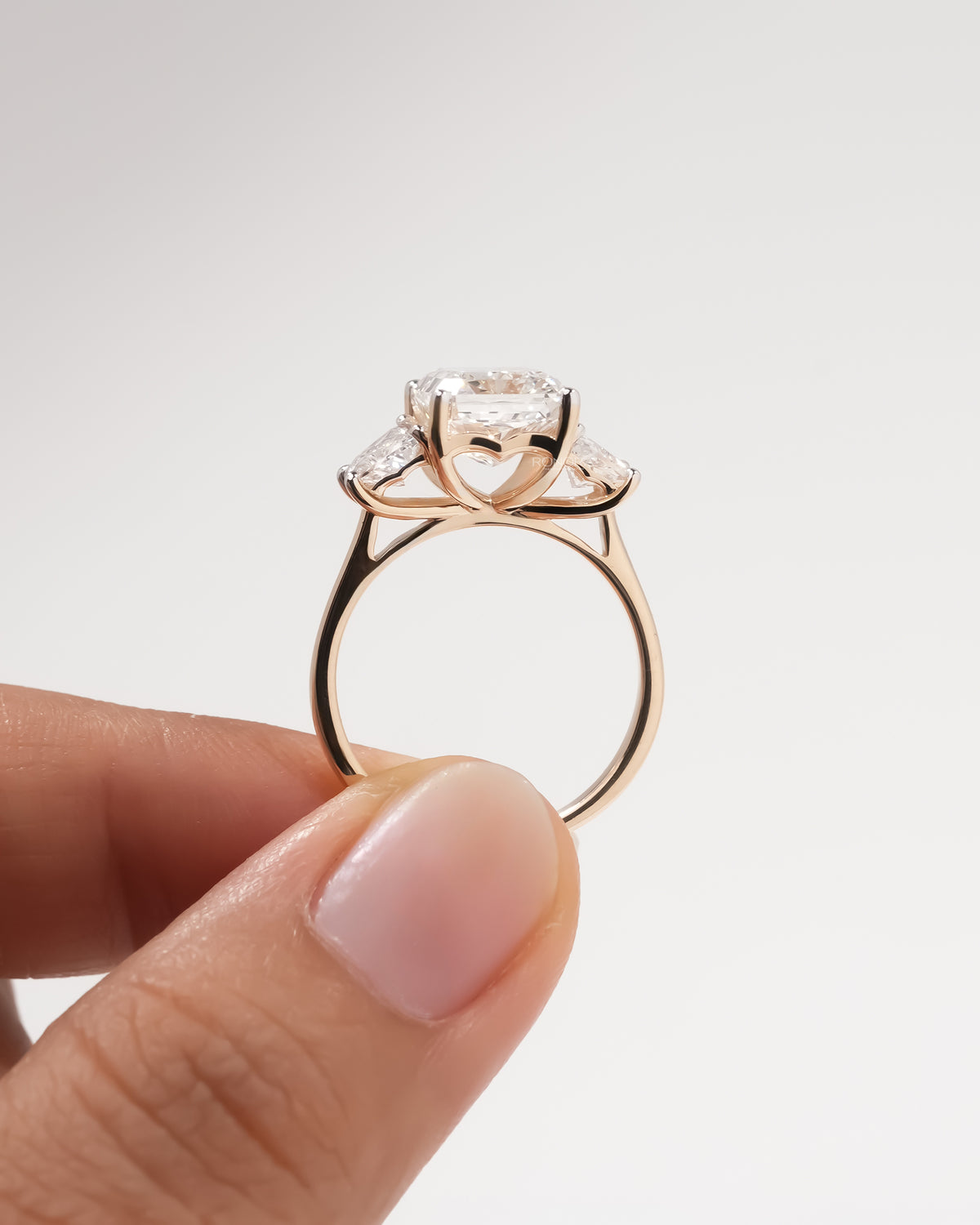 Back view of a rose gold radiant cut lab grown diamond three stone ring held up between two fingers